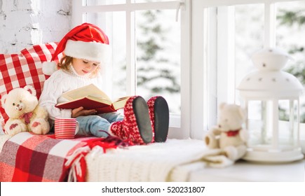 Happy Child Girl Reading A Book While Sitting At A Winter Window Christmas