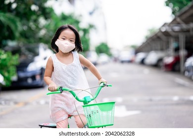 Happy Child Girl Rides A Bicycle On The Road Around Her House In The Evening Time. Kid Wearing Cloth Face Mask With A Filter Filling Inside To Protect Spread Coronavirus (covid-19) And Pm2.5.