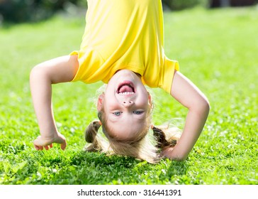 Happy Child Girl Standing Upside Down On Her Head On Grass In Summer