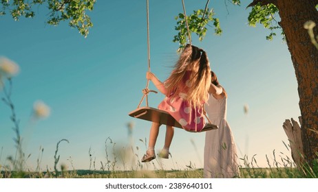 Happy child girl is swinging on swing in park at sunset. Daughter, mama play with wooden swings on tree. Happy family. Kid flies on swing. Children entertainment in park with family. Child dream fly. - Powered by Shutterstock