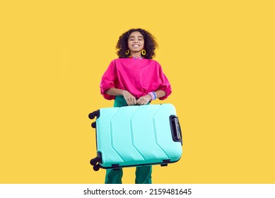 Happy Child Going On Summer Vacation. Black Kid With Suitcase Standing Isolated On Yellow Background. Beautiful African American Girl In Trendy Casual Outfit Carrying Her Heavy Turquoise Travel Bag