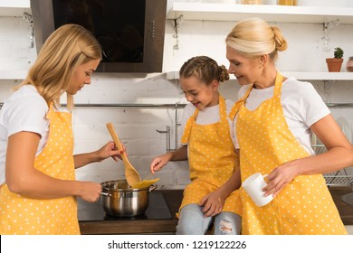 Happy Child With Grandmother In Aprons Looking At Young Mother Cooking Pasta In Kitchen