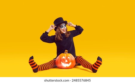 Happy child having fun on Halloween. Funny little girl with Catrina skull make up in top hat and striped orange black stockings sitting on floor with Jack o lantern pumpkin on yellow studio background - Powered by Shutterstock