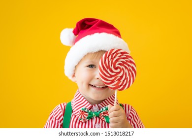 Happy child holding lollipop in hand against yellow background. Portrait of funny kid dressed Santa Claus hat. Christmas holiday concept