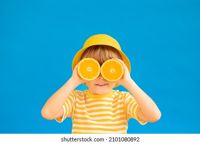 Happy Child Holding Slices Of Orange Fruit Like Sunglasses. Kid Wearing Striped Yellow T-shirt Against Blue Paper Background. Healthy Eating And Summer Vacation Concept