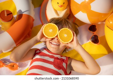 Happy Child Holding Slices Of Orange Fruit Like Sunglasses. Kid Wearing Striped T-shirt Lying On Beach Towel. Healthy Eating And Summer Vacation Concept