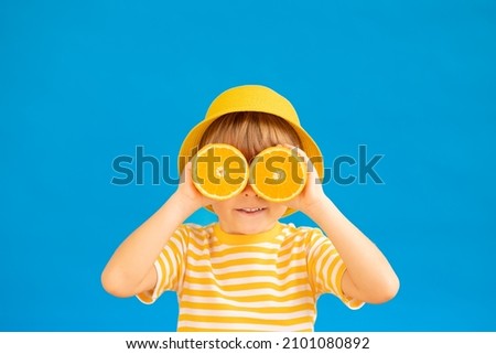 Happy child holding slices of orange fruit like sunglasses. Kid wearing striped yellow t-shirt against blue paper background. Healthy eating and summer vacation concept