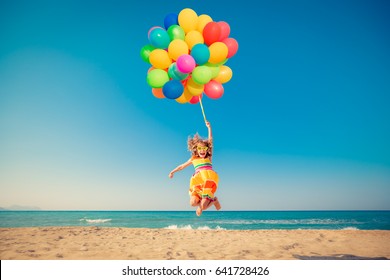 Happy child jumping with colorful balloons on sandy beach. Portrait of funny girl against blue sea and sky background. Active kid having fun on summer vacation. Freedom and imagination concept