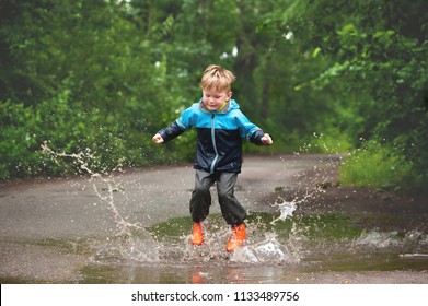 Happy Child Jumping In Puddle In Waterproof Coat. A Boy Have Fun In Rain In A Bright Raincoat. Kid Playing In Mud With Splash In Autumn Park. Outdoor Fun In The Rain Weather