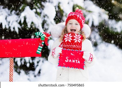 Happy Child In Knitted Reindeer Hat And Scarf Holding Letter To Santa With Christmas Presents Wish List At Red Mail Box In Snow Under Xmas Tree In Winter Forest. Kids Sending Post To North Pole.