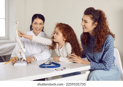 Happy child learning about human skeleton during visit to doctor. Mom and little girl sitting at table in pediatrician's office and looking at orthopedic spine model. Kids, anatomy, medicine concept - Powered by Shutterstock