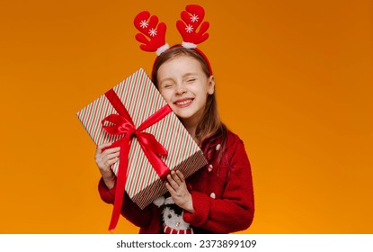 Happy child with the long-awaited gift. A girl in a red Christmas sweater presses a gift box to her cheek. The girl closed her eyes with pleasure that her dream had come true.
