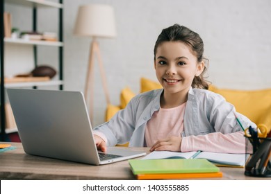 happy child looking at camera while sitting at desk and using laptop at home