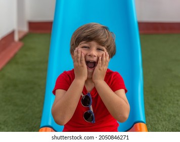 Happy Child On A Blue Slide In The Garden Of A House On Artificial Grass