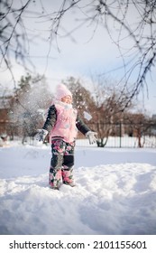 Happy child on winter walk. Little girl in warm suit plays in backyard and enjoys sunny day in winter, she stands on snowdrift and plays snowballs.