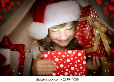 Happy child opening Christmas gift box. Funny baby dressed in Santa Claus hat in bedroom. Portrait of smiling kid at home. Xmas holiday concept