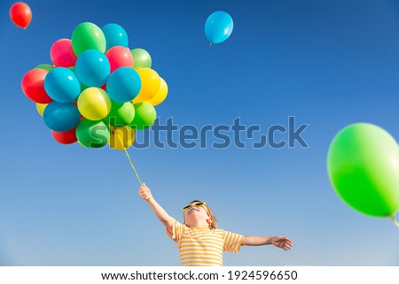 Happy child playing with bright multicolor balloons outdoor. Kid having fun in green spring field against blue sky background. Healthy and active lifestyle concept
