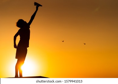 Happy Child Is Playing With Paper Origami Plane At Orange Sunset. Vocation With Children Outdoors. Full Size Silhuette Against Sunset.