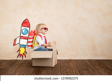 Happy Child Playing With Toy Rocket At Home. Funny Kid Driving Cardboard Box Indoor. Success And Win Concept