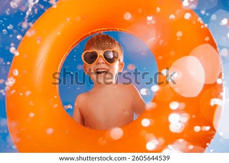 Happy child playing in waterpark. Kid having fun on summer vacation. Boy jumping into swimming pool with splashes of water. Funny child holding orange ring against blue sky background