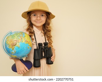 Happy Child Plays The Discoverer. Unique Child. Travel, Adventure, Vacation And Discovery Concept. A Kid In A Traveler's Hat With Binoculars And A Globe In His Hands On The Beige Background. 