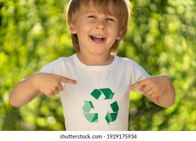 Happy Child Points Fingers At Recycle Sign On T-shirt. Funny Kid Against Spring Green Background. Ecology And Earth Day Concept. 