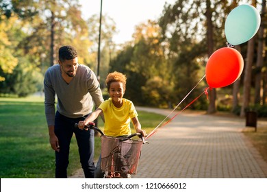 Happy Child Riding A Bicycle. Father Teaching His Daughter How To Ride A Bike. Birthday Present, Balloons Flying.