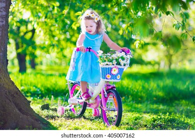 Happy Child Riding A Bike. Cute Kid Biking Outdoors. Little Girl In A Blue Dress On A Pink Bicycle With Daisy Flowers In A Basket. Healthy Preschool Children Summer Activity. Kids Playing Outside.