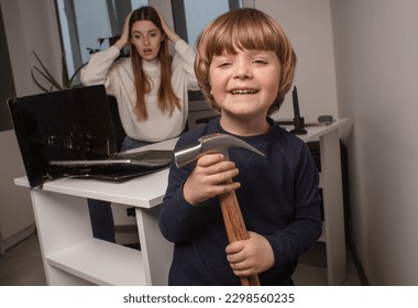 Happy child smashes mom's work laptop to spend more time together. The toddler laughs because he broke his mother's computer. 
The naughty son broke the shocked mother's computer with a hammer.
