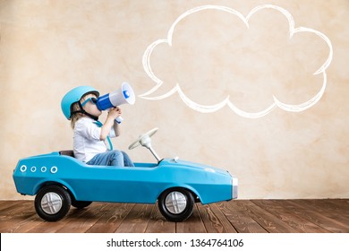 Happy Child Speaking To Megaphone At Home. Funny Kid Driving Toy Car Indoor. Success And Win Concept