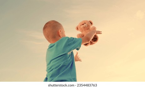 Happy child with teddy bear outdoors on summer day. Little boy throws up and catches bear toy against background of summer nature at sunset. Family weekend in nature. Concept of child's dream to fly - Powered by Shutterstock