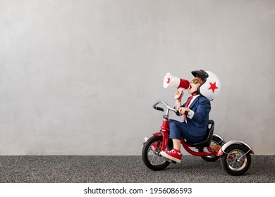 Happy child wearing suit riding bicycle against grey concrete wall background. Funny kid shouting through loudspeaker. Childhood dreams and business idea concept