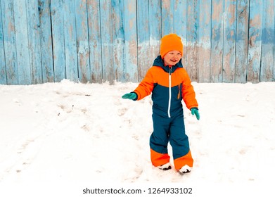 Happy Child In Winter Warm Clothes Run Through Snow On Eve In Village Yard. Fence With Peeling Paint, Rustic Fun, Joy Of  Brothers. Children Are Happy On Vacation In  Country