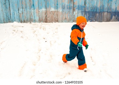 Happy Child In Winter Warm Clothes Run Through Snow On Eve In Village Yard. Fence With Peeling Paint, Rustic Fun, Joy Of  Brothers. Children Are Happy On Vacation In  Country