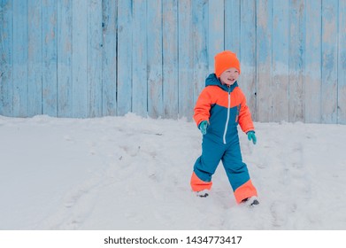 Happy Child In Winter Warm Clothes Run Through Snow On Eve In Village Yard. Fence With Peeling Paint, Rustic Fun, The Joy. Child Are Happy On Vacation In The Country