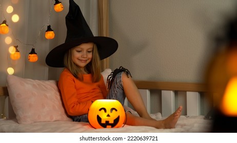 Happy Child In Witch Black Hat Plays With Spider In Dark With Orange Pumpkin Lantern At Home. Halloween