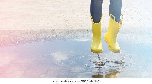 Happy child in yellow wellington boot jumps in a puddle of water after rain. Cheerful baby on a summer sunny day. Outdoor games. Copy space.