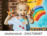 Happy childhood. Close-up of a funny little girl, stained with colorful paints and with brushes in her hair, shouting for joy. An easel with a child’s drawing is in the background. Kids imagination.