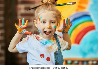 Happy childhood. Close-up of a funny little girl, stained with colorful paints and with brushes in her hair, shouting for joy. An easel with a child’s drawing is in the background. Kids imagination. - Powered by Shutterstock