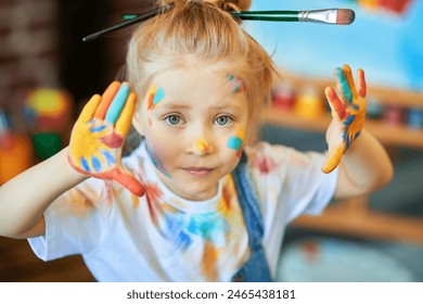 Happy childhood. Close-up of a funny little girl, stained with colorful paints and with brushes in her hair, smiling for joy. An easel with a child’s drawing is in the background. Kids imagination.