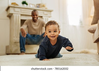 Happy childhood. Happy dark-haired afro-american man laughing and watching his cheerful young son crawling on the floor