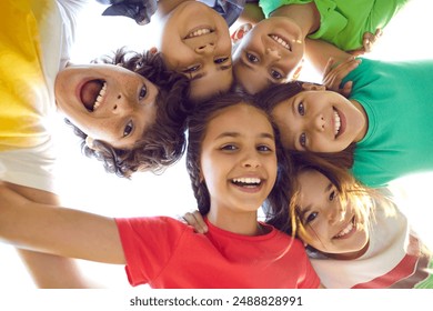 Happy childhood. Faces of funny joyful preteen friends who stand in circle and look down at camera. Bottom view of cute boys and girls in colorful summer casual clothes hugging and laughing outdoors.