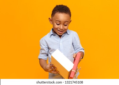 Happy Childhood, Joy And Celebration Concept. Cheerful Fashionable Dark Skinned Boy In Blue Shirt Opening Box With Gift Received From His Friend. Cute Black Child Overjoyed With Birthday Present