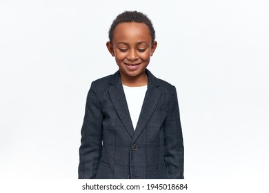 Happy Childhood, Joy And Positive Emotions Concept. Isolated Image Of Adorable Cheerful Black Little Boy In Formal Clothing Looking Down With Shy Smile. Timid Dark Skinned Child Smiling In Studio