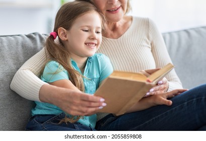 Happy Childhood. Loving Grandmother Teaching Granddaughter Holding Book Sitting On Sofa, Grandma Baby Sitter Embracing Kid Girl Reading Fairytale To Cute Child, Nanny Granny Telling Story