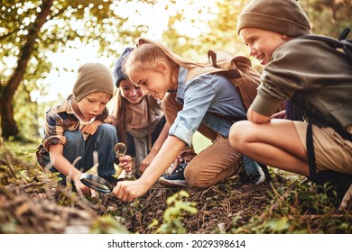 Happy children boys and girls in casual clothes with backpacks making bonfire with magnifying glass together in green forest during school camping activity on sunny day, smiling kids exploring nature
