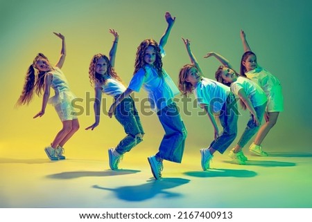 Happy children dancing. Group of children, little girls in sportive casual style clothes dancing in choreography class isolated on green background in yellow neon light. Concept of music, fashion, art