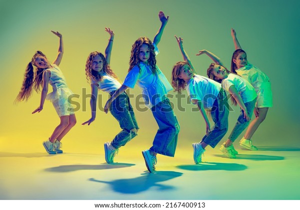 Happy children dancing. Group of children, little\
girls in sportive casual style clothes dancing in choreography\
class isolated on green background in yellow neon light. Concept of\
music, fashion, art