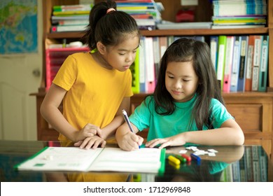 Happy Children Doing Homework.Primary Students Having Lesson Together.Cheerful Asian Pupils.Homeschooling.Kid Writing On Workbook.Map And Shelves At Background.Files And Crayons.