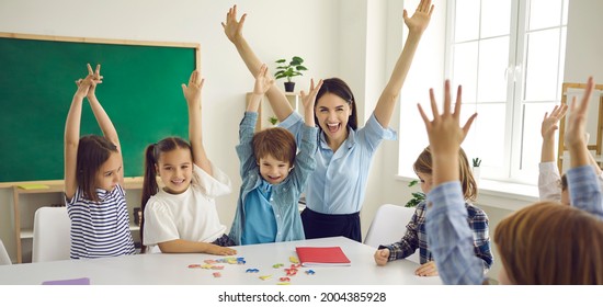 Happy children and educator raising hands up sitting at table in modern classroom. Website header with group portrait of cheerful school teacher and first grade students having fun activities in class - Powered by Shutterstock